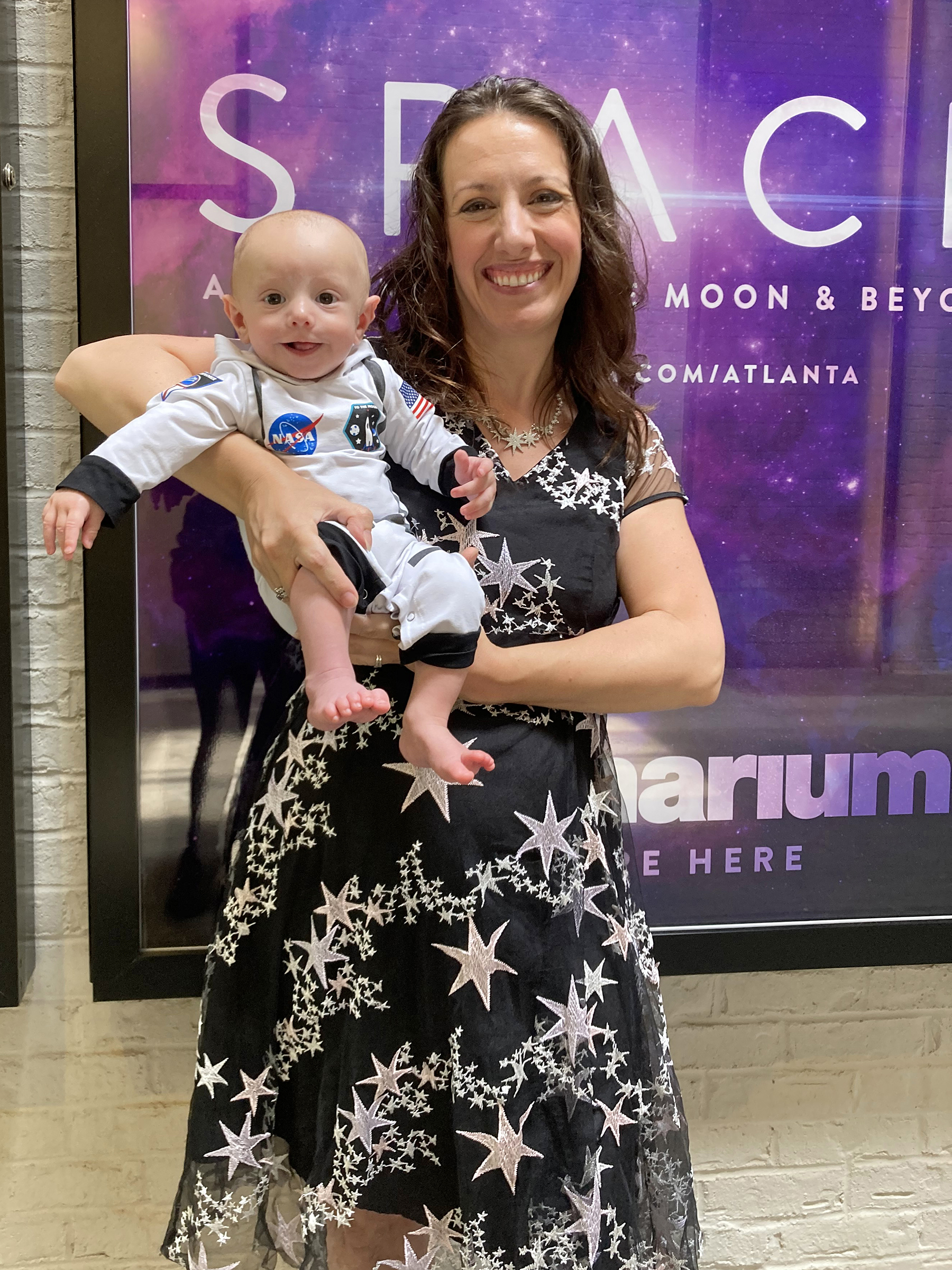 Laura Forczyk wearing black dress with stars holding a smiling baby in a NASA astronaut onesie standing in front of purple space poster
