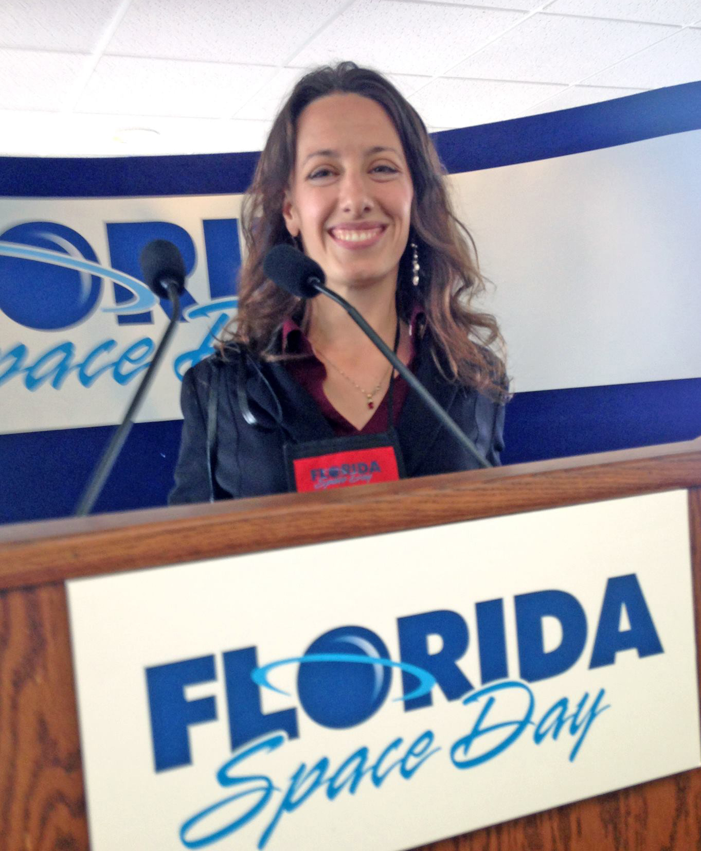 Laura Forcyk smiling for photo standing behind a microphone attached to a speaking platform with the Florida Space Day logo on the front